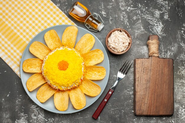 Vista superior de la ensalada en el lado izquierdo con una servilleta amarilla debajo con una botella de aceite de tenedor de queso y una tabla de cortar en el lado de la mesa gris oscuro