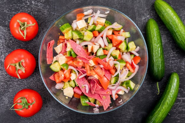 Vista superior de ensalada de pepino picado en un recipiente de vidrio con tomate y verduras frescas en piedra negra