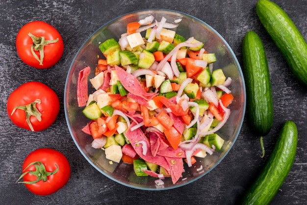 Vista superior de ensalada de pepino picado en un recipiente de vidrio con tomate y verduras frescas en piedra negra
