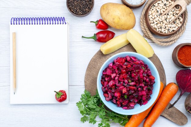 Vista superior de ensalada de remolacha fresca con verduras en rodajas junto con frijoles crudos, zanahorias, patatas, bloc de notas en el escritorio de luz, comida comida ensalada fresca