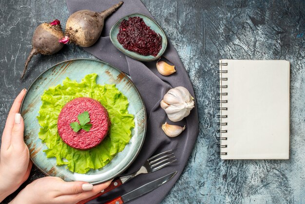 Vista superior de la ensalada de remolacha en el plato en manos de la mujer remolacha con ajo remolacha rallada en un tazón pequeño, tenedor y cuchillo, bloc de notas con mantón morado en la mesa gris