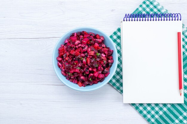 Vista superior de la ensalada de remolacha en rodajas con verduras dentro de la placa azul con el bloc de notas en el escritorio de luz, ensalada de alimentos vegetales salud de la comida