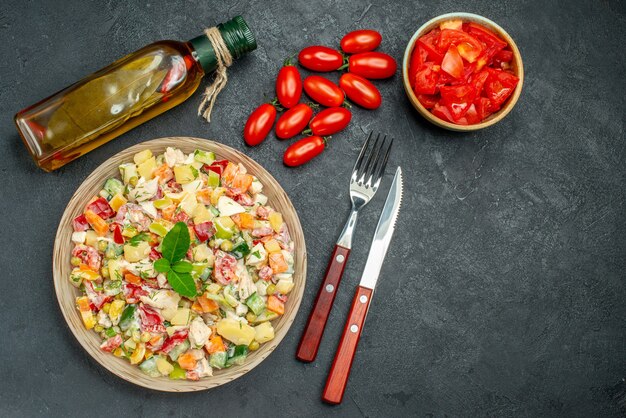 Vista superior de ensalada de verduras con botella de aceite de tomate y cubiertos en el lado sobre fondo gris oscuro