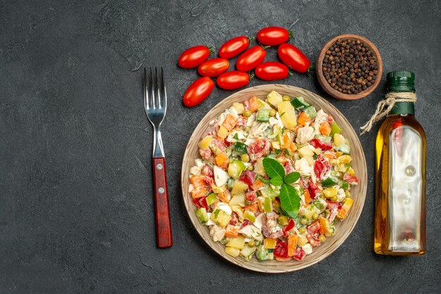 Vista superior de la ensalada de verduras con una botella marcada de aceite de tomates pimientos y un tenedor en el costado sobre fondo oscuro