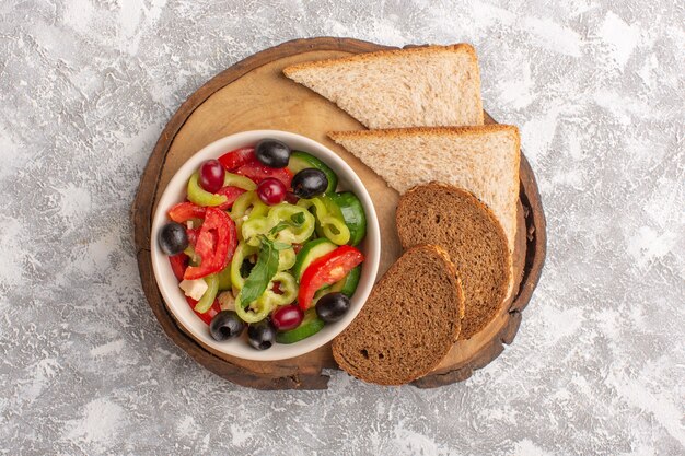 Vista superior Ensalada de verduras frescas con pepinos en rodajas, tomates, aceitunas y queso blanco dentro de la placa con pan de molde en la comida de ensalada de comida vegetal de escritorio gris