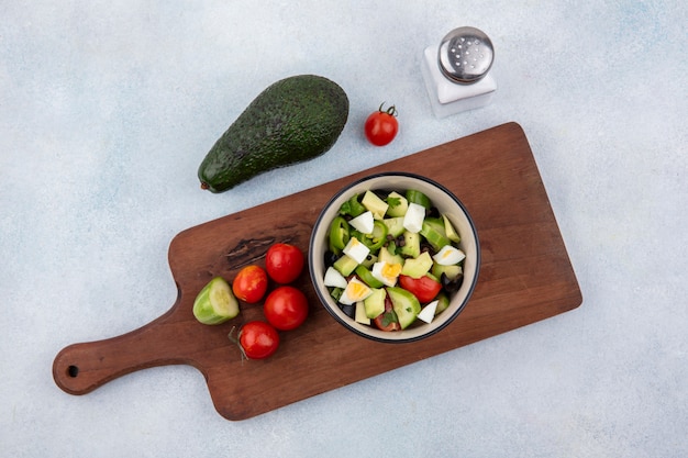 Vista superior de ensalada de verduras frescas que incluye tomate pimiento pepino en un recipiente sobre tablero de cocina de madera con aguacate y salero en blanco