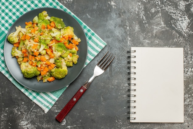 Vista superior de ensalada de verduras frescas y saludables en una toalla despojada verde y un cuaderno en la mesa gris