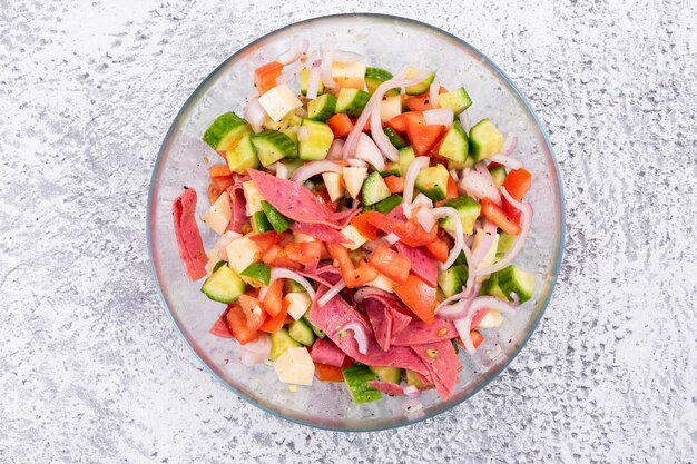 Vista superior ensalada de verduras picadas en un recipiente de vidrio con tomate cebolla pepino en piedra blanca