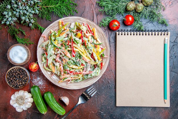 Vista superior ensalada de verduras en plato tenedor tomates ramas de pino pepinos ajo lápiz en el bloc de notas en la mesa de color rojo oscuro