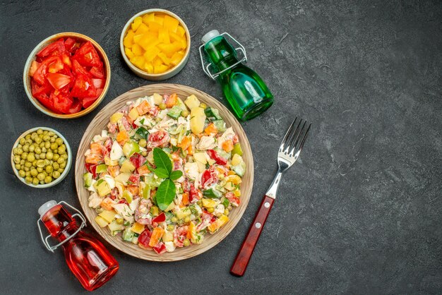 Vista superior de la ensalada de verduras con tenedor botellas de aceite y vinagre y tazones de verduras en el lado de la mesa gris oscuro