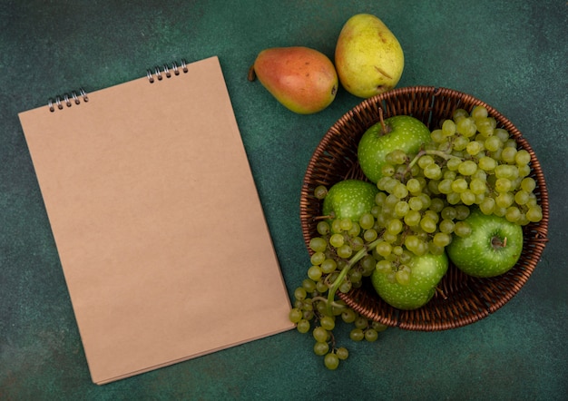 Vista superior del espacio de copia beige Bloc de notas con uvas verdes y manzanas en una canasta de peras sobre un fondo verde