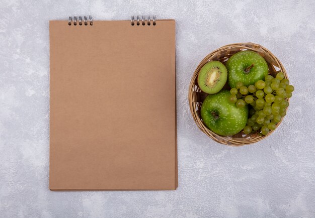 Vista superior espacio de copia manzanas verdes con uvas verdes y cuña de kiwi en canasta con bloc de notas marrón sobre fondo blanco.