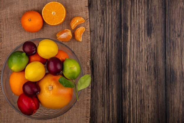 Vista superior espacio de copia mezcla de frutas limones limas ciruela melocotón y naranja en un jarrón sobre una servilleta beige sobre un fondo de madera