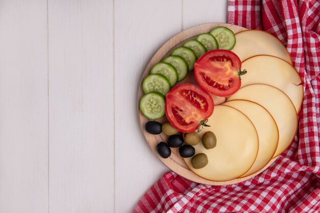 Vista superior del espacio de copia de queso ahumado con tomates, pepinos y aceitunas en un soporte con una toalla a cuadros roja sobre un fondo blanco.