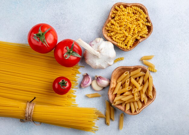 Vista superior de espaguetis crudos con pasta cruda en tazones con ajo y tomates sobre una superficie gris