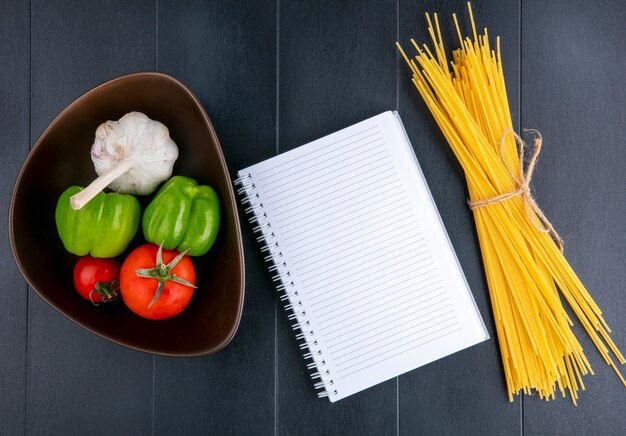 Vista superior de espaguetis crudos con tomates, ajo y pimiento en un recipiente y bloc de notas sobre una superficie negra