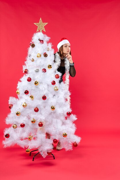 Vista superior del estado de ánimo de año nuevo con hermosa chica en un vestido negro con sombrero de santa claus escondido detrás del árbol de navidad