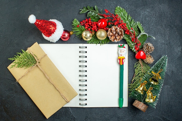 Vista superior del estado de ánimo navideño con ramas de abeto, sombrero de santa claus, árbol de Navidad, caja de regalo en el cuaderno sobre fondo oscuro