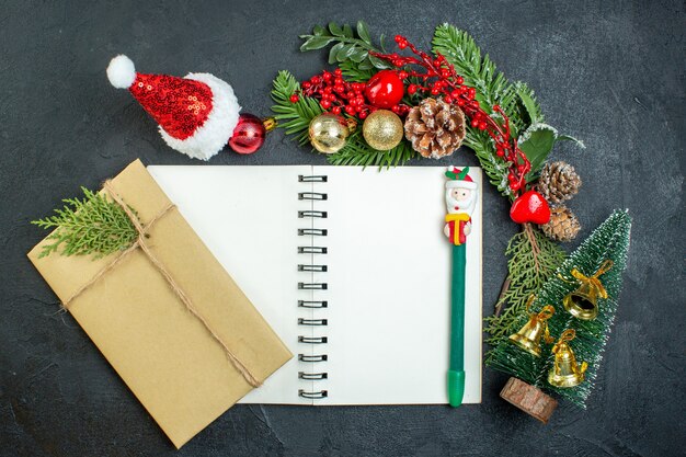 Vista superior del estado de ánimo navideño con ramas de abeto, sombrero de santa claus, árbol de Navidad, caja de regalo en el cuaderno sobre fondo oscuro