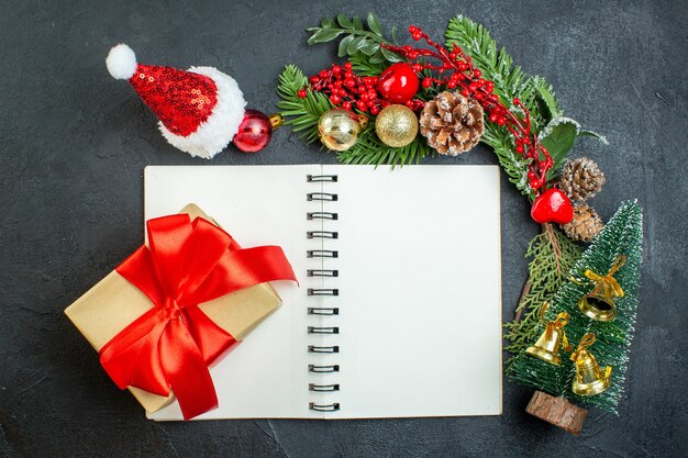 Vista superior del estado de ánimo navideño con ramas de abeto, sombrero de santa claus, árbol de Navidad, caja de regalo en el cuaderno sobre fondo oscuro