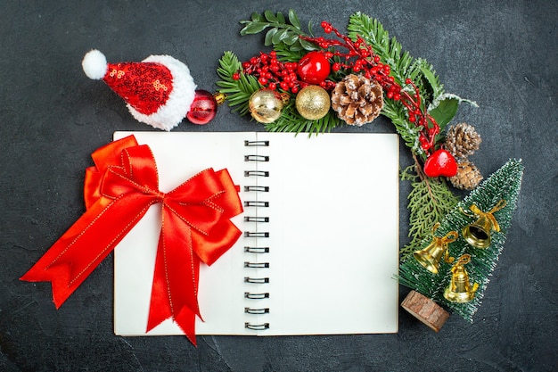 Vista superior del estado de ánimo navideño con ramas de abeto, sombrero de santa claus, árbol de navidad, cinta roja en el cuaderno sobre fondo oscuro