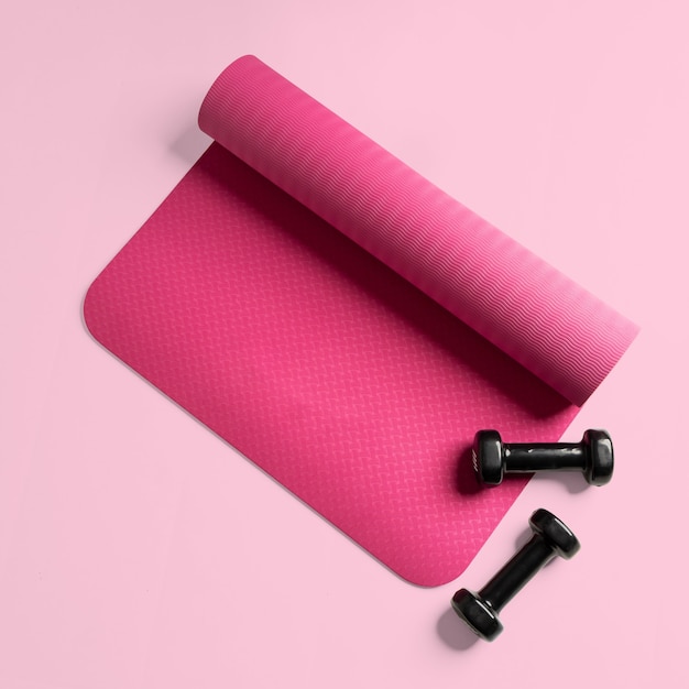 Vista superior de una estera de fitness rosa y dos pesas negras aisladas sobre una superficie rosa