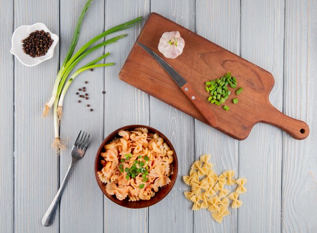 Vista superior de farfalle pasta con cebolla verde picada en un tazón tabla de cortar de madera con cuchillo y ajo sobre fondo rústico