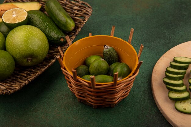 Vista superior de feijoas en un balde con rodajas de pepino picadas en una tabla de cocina de madera con manzanas verdes, aguacates, pepino en una bandeja de mimbre sobre un fondo verde