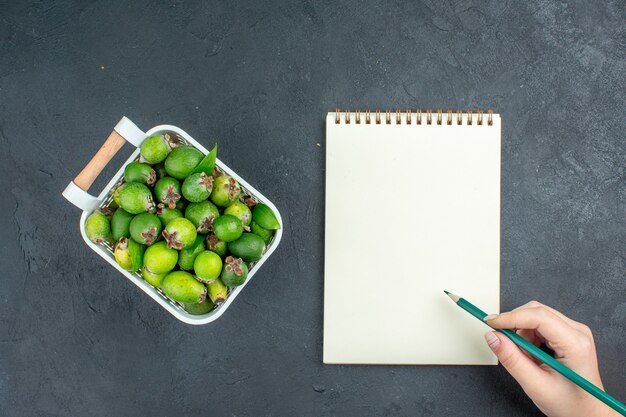 Vista superior feijoas frescas en canasta de plástico verde pwncil en bloc de notas de mano femenina sobre superficie oscura