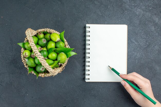 Vista superior feijoas frescas en lápiz de cuaderno de canasta en mano femenina sobre superficie oscura