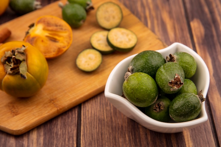 Foto gratuito vista superior de feijoas frescas en un recipiente con rodajas de feijoas con frutos de caqui en una tabla de cocina de madera en una pared de madera