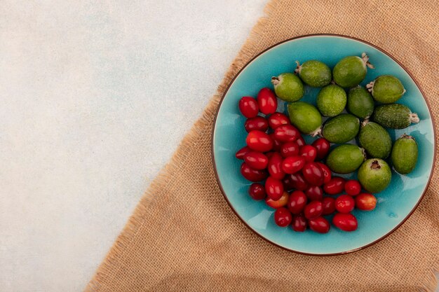 Vista superior de feijoas maduras con cerezas de cornalina en un plato azul sobre una tela de saco sobre una superficie gris con espacio de copia