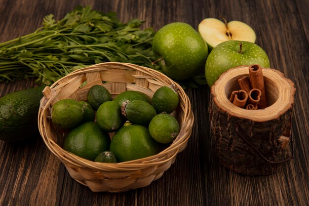 Vista superior de feijoas maduras frescas en un balde con ramas de canela en un frasco de madera con manzanas y aguacates aislado en una pared de madera