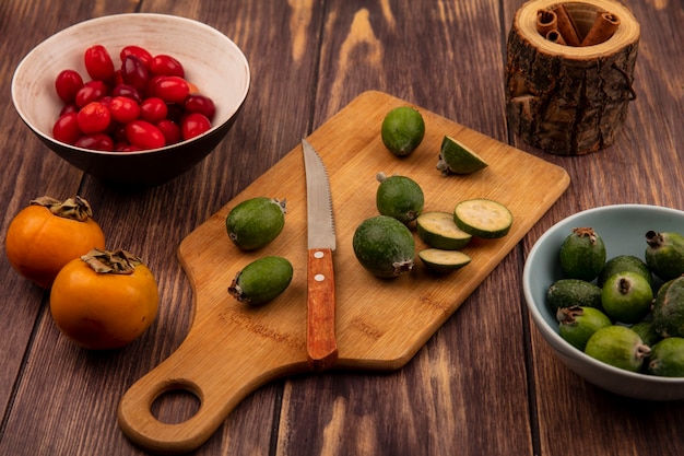 Vista superior de feijoas en una tabla de cocina de madera con cuchillo con cerezas de cornalina en un recipiente con ramas de canela sobre un fondo de madera