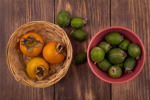 Foto gratuita vista superior de feijoas verdes frescas en un recipiente con frutos de caqui en un balde sobre una superficie de madera