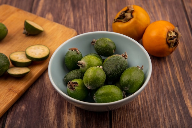 Vista superior de feijoas verdes en un recipiente con rodajas de feijoas en una tabla de cocina de madera con frutos de caqui aislado en una pared de madera