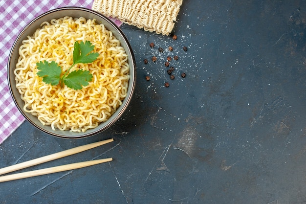 Vista superior de fideos ramen asiáticos con cilantro en un tazón sobre mantel a cuadros blanco rosa palillos fideos crudos en la mesa oscura