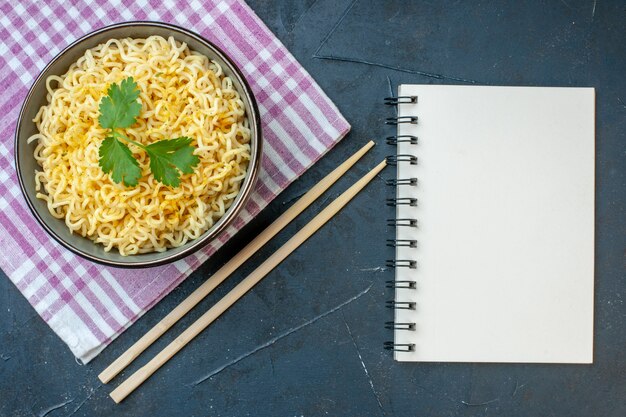 Vista superior de fideos ramen asiáticos en un tazón en palillos de bloc de notas de servilleta a cuadros rosa blanco en la mesa oscura