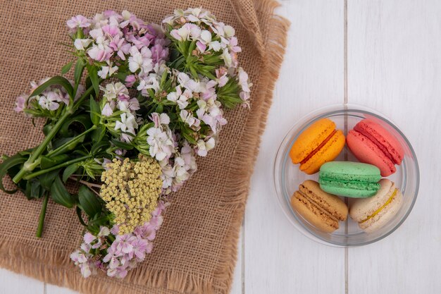 Vista superior de flores en una servilleta beige con macarons de colores en un frasco sobre una superficie blanca