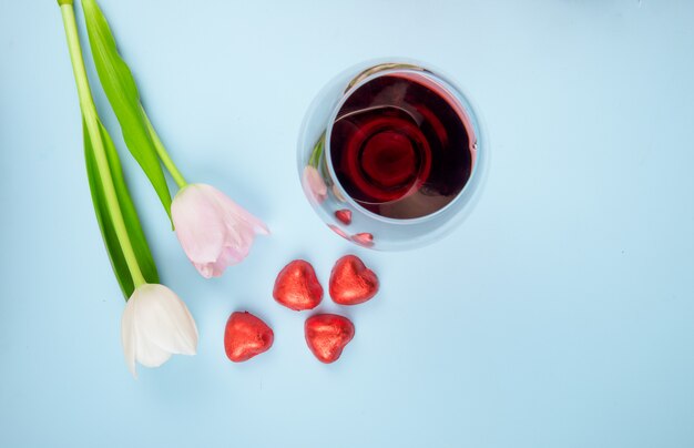 Vista superior de flores de tulipán de color blanco y rosa con caramelos dispersos en forma de corazón en papel rojo y una copa de vino en la mesa azul