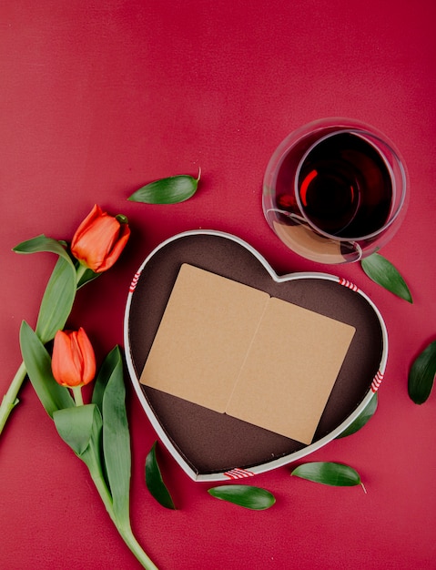 Vista superior de flores de tulipán de color rojo con caja de regalo en forma de corazón con una postal abierta y una copa de vino tinto sobre fondo rojo.