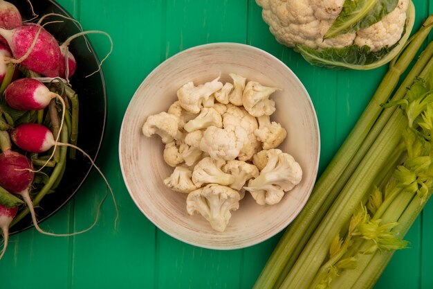 Vista superior de floretes de coliflor fresca en un recipiente con rábanos en un recipiente con coliflor entera y apio aislado en una pared de madera verde