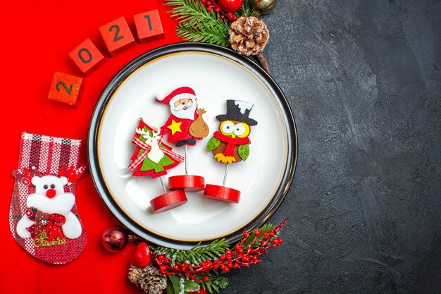 Vista superior del fondo de año nuevo con accesorios de decoración de plato de cena ramas de abeto y números calcetín de Navidad en una servilleta roja sobre una mesa negra