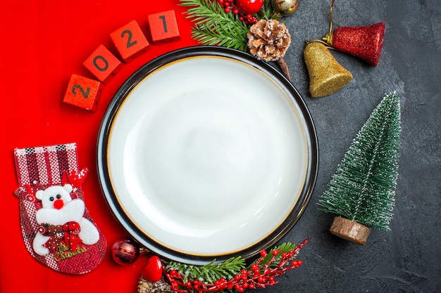 Vista superior del fondo de año nuevo con accesorios de decoración de plato de cena ramas de abeto y números en una servilleta roja junto al árbol de Navidad en una mesa negra