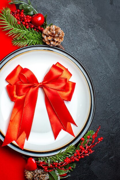 Vista superior del fondo de año nuevo con cinta roja en el plato de cena cubiertos set accesorios de decoración ramas de abeto junto a un regalo en una servilleta roja sobre una mesa negra