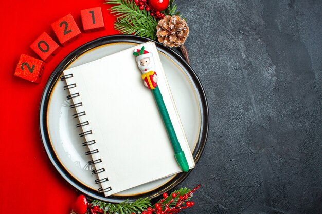 Vista superior del fondo de año nuevo con cuaderno de espiral en accesorios de decoración de plato de cena ramas de abeto y números en una servilleta roja sobre una mesa negra