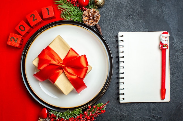 Foto gratuita vista superior del fondo de año nuevo con regalo en accesorios de decoración de plato de cena ramas de abeto y números en una servilleta roja y cuaderno con lápiz sobre una mesa negra