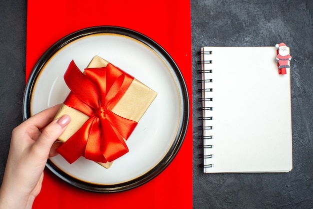 Foto gratuita vista superior del fondo de comida nacional christmal con mano sosteniendo platos vacíos con cinta roja en forma de arco en una servilleta roja y cuaderno sobre mesa negra