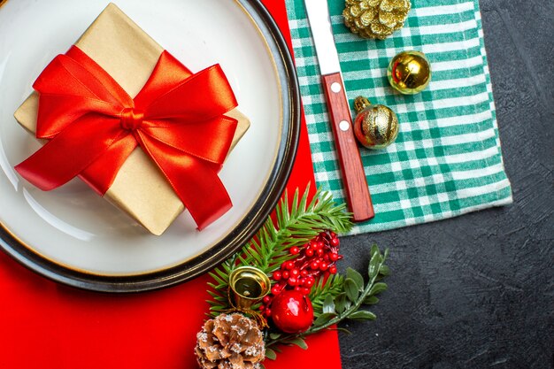 Vista superior del fondo de comida nacional Christmal con regalo con cinta roja en forma de arco en platos vacíos cubiertos accesorios de decoración en toalla verde despojada