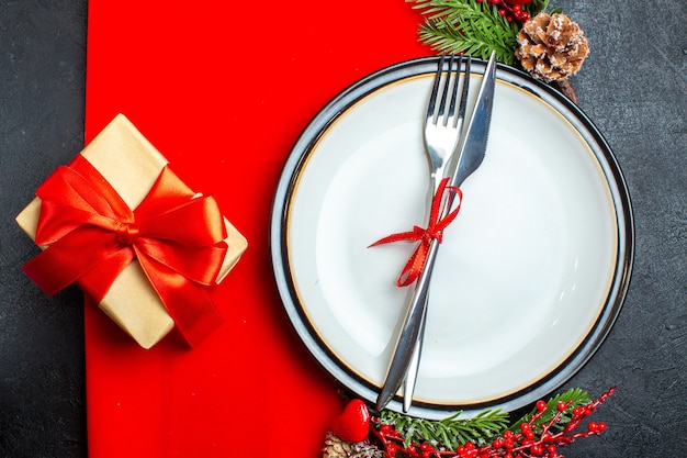 Vista superior del fondo de Navidad con cubiertos con cinta roja en un plato de cena accesorios de decoración ramas de abeto junto a un regalo en una servilleta roja